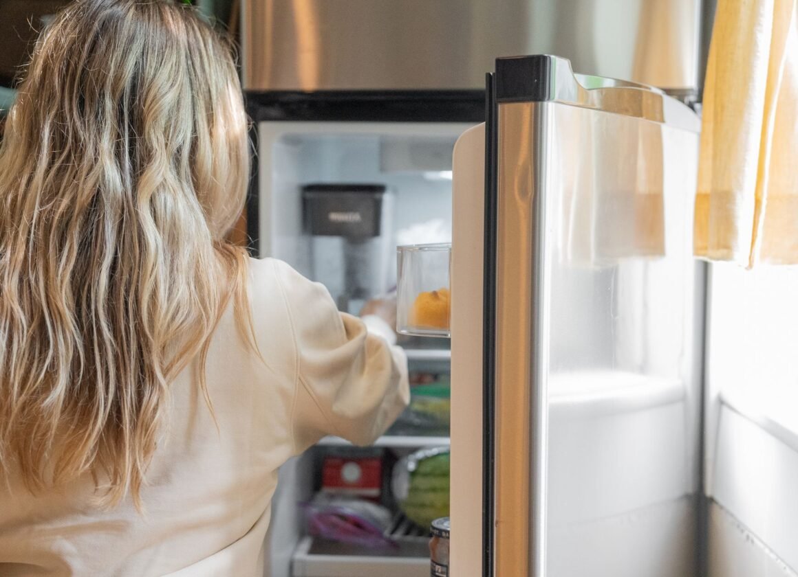 Back view of a blonde woman retrieving food from a refrigerator in a bright kitchen.